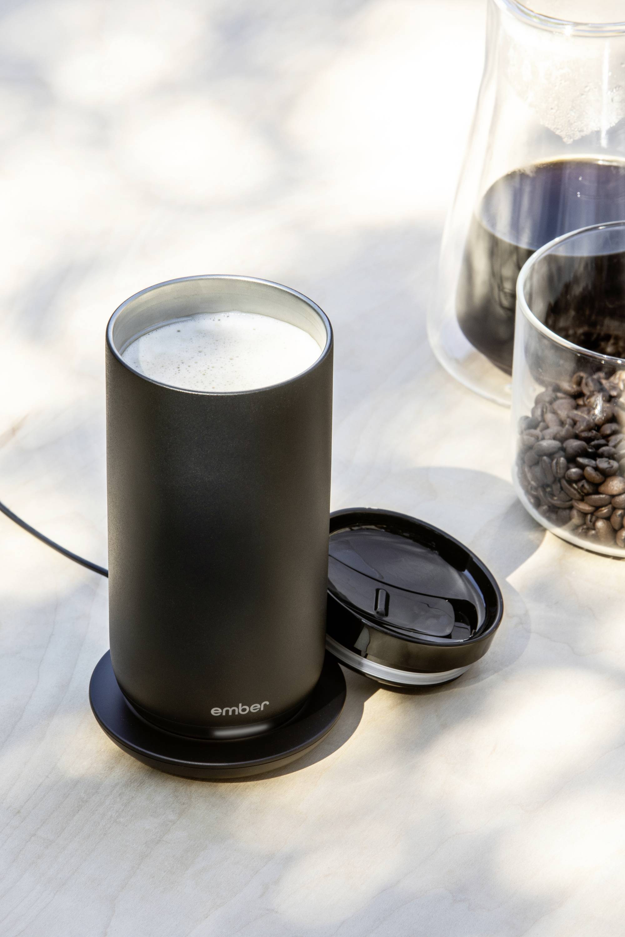 A black thermal mug on a desk, filled with a beverage, alongside a bowl of coffee beans and a coffee pot.