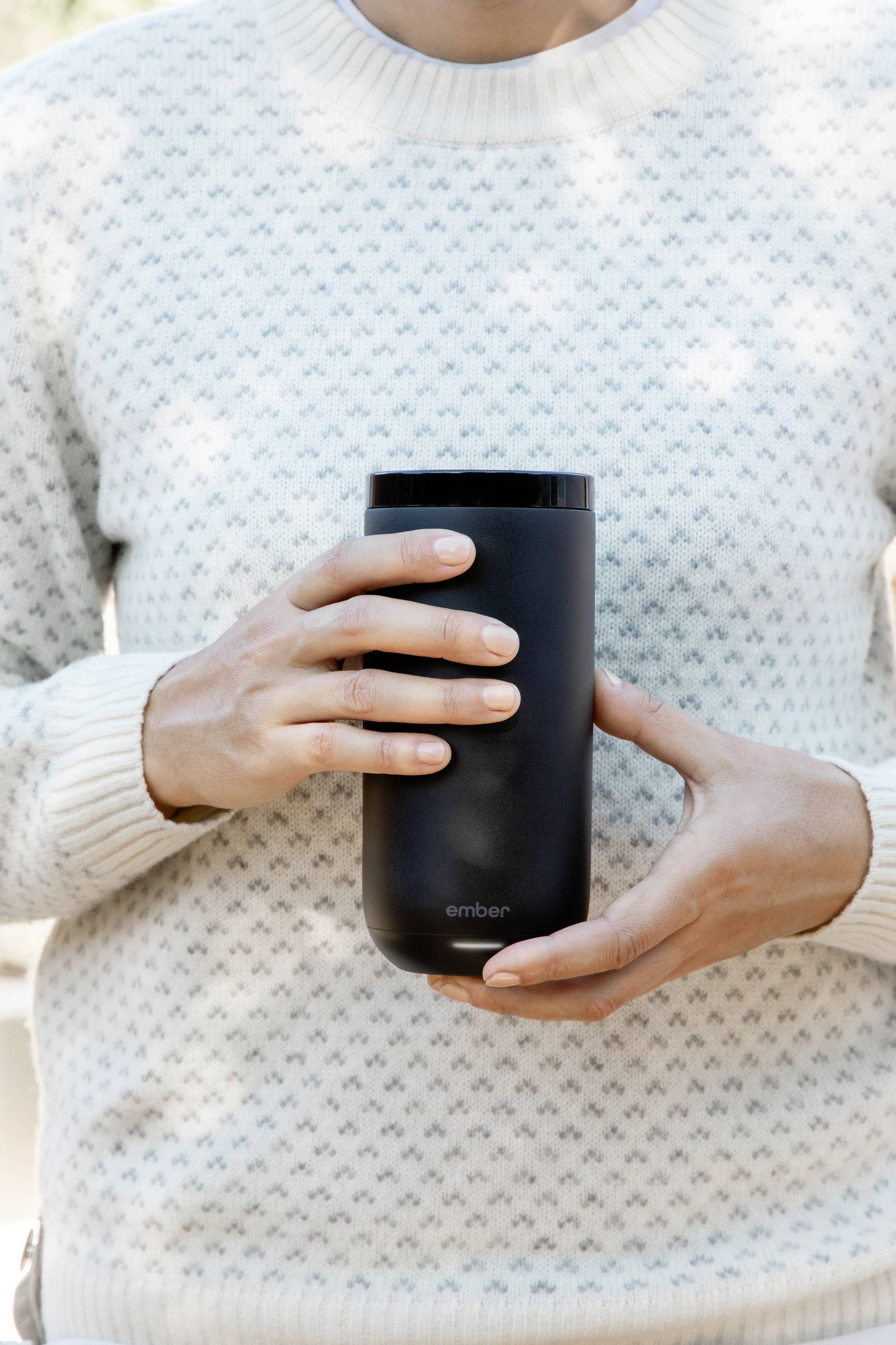 A person is holding a black thermal mug in both hands. Background blurred, focus on the mug.