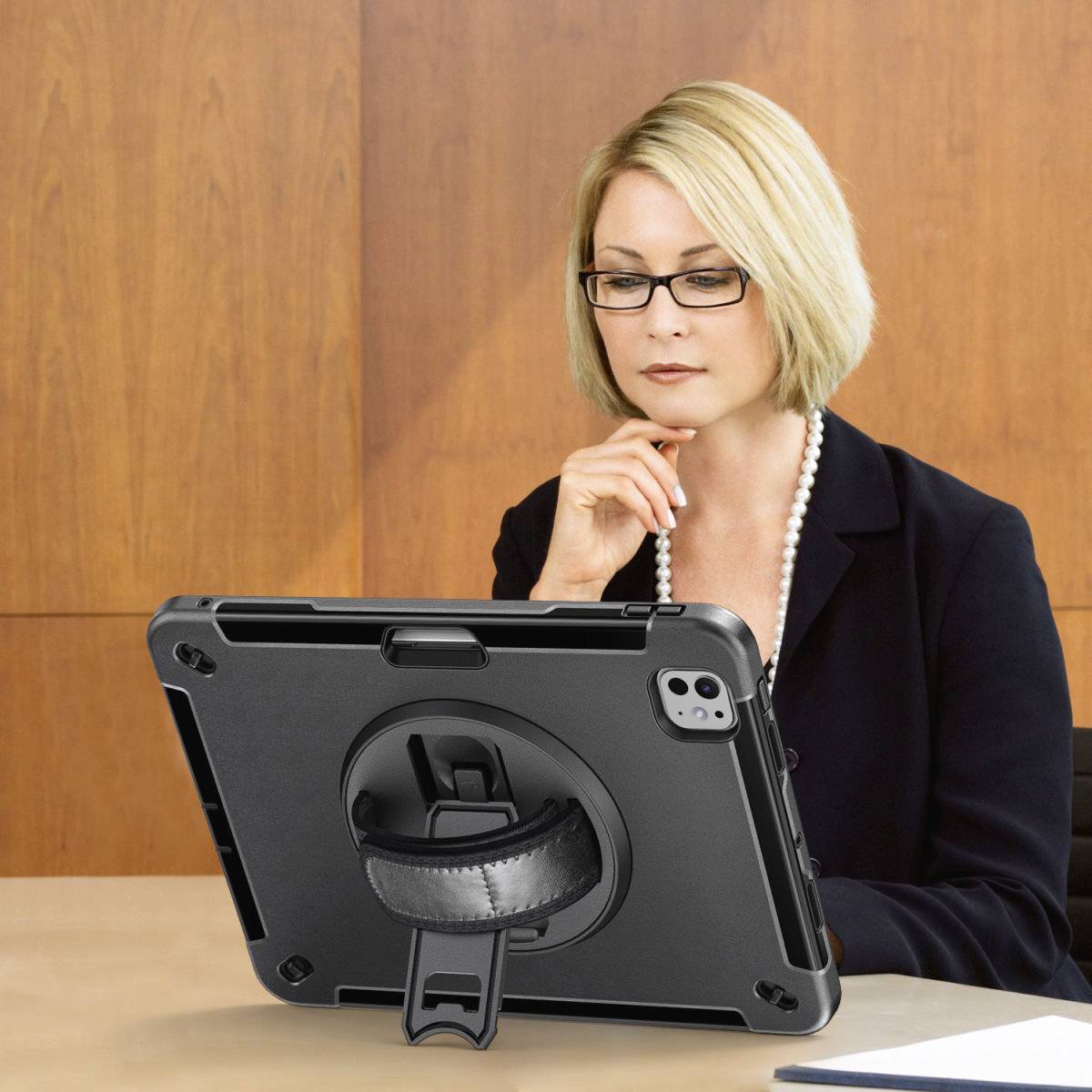 A person sits at a table, contemplating a tablet thoughtfully. They are wearing glasses and business attire.