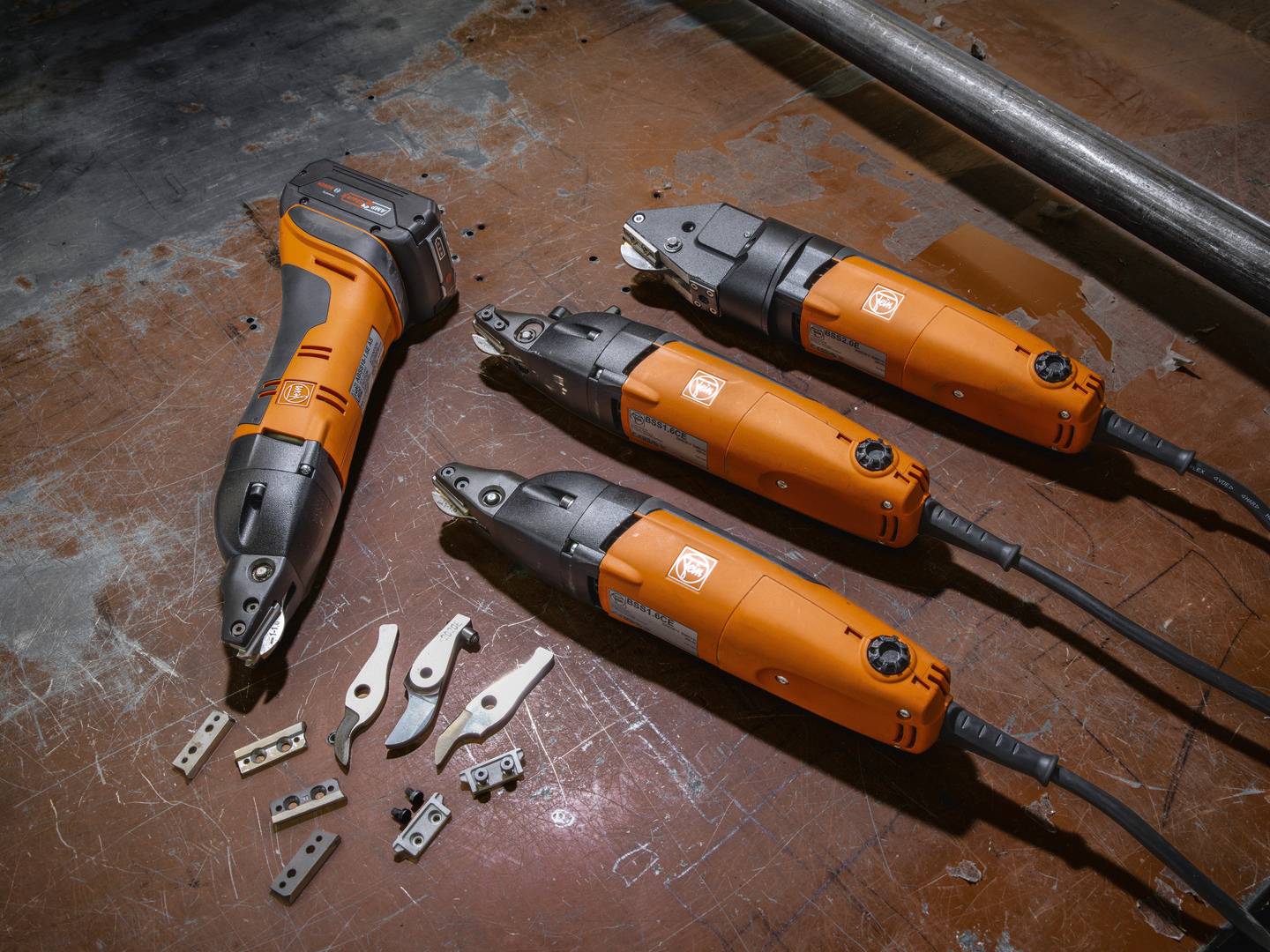 Four orange electric power tools with blade attachments on a table, surrounded by several loose blades and accessory parts.