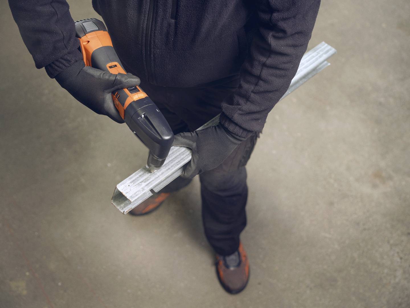 A person is working with a drill on a piece of metal, wearing black workwear and gloves.