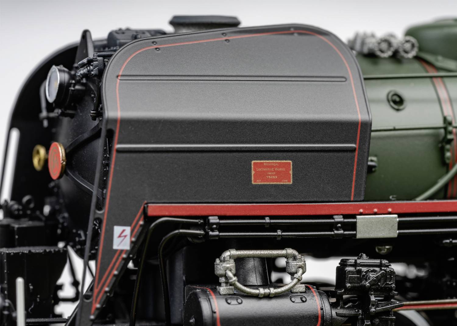 Close-up of part of a steam locomotive, showing technical details and a red stripe on black metal. An electricity symbol is on the left.