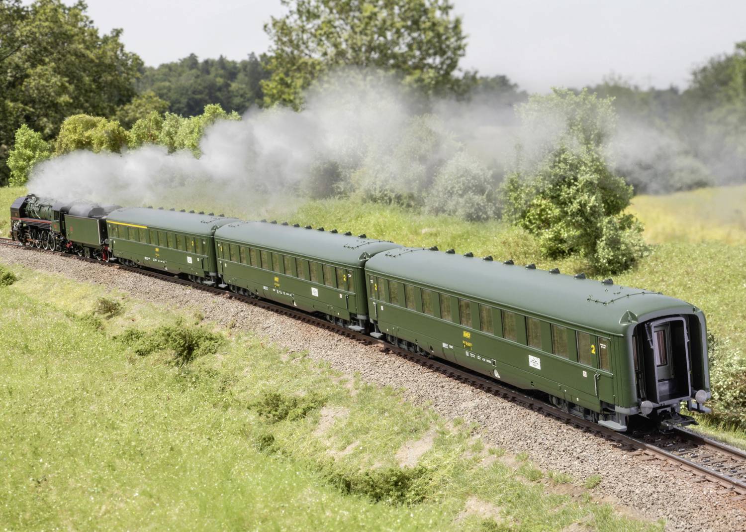 Train with green carriages travelling through a verdant landscape; plume of smoke reveals steam locomotive. Trees in the background.