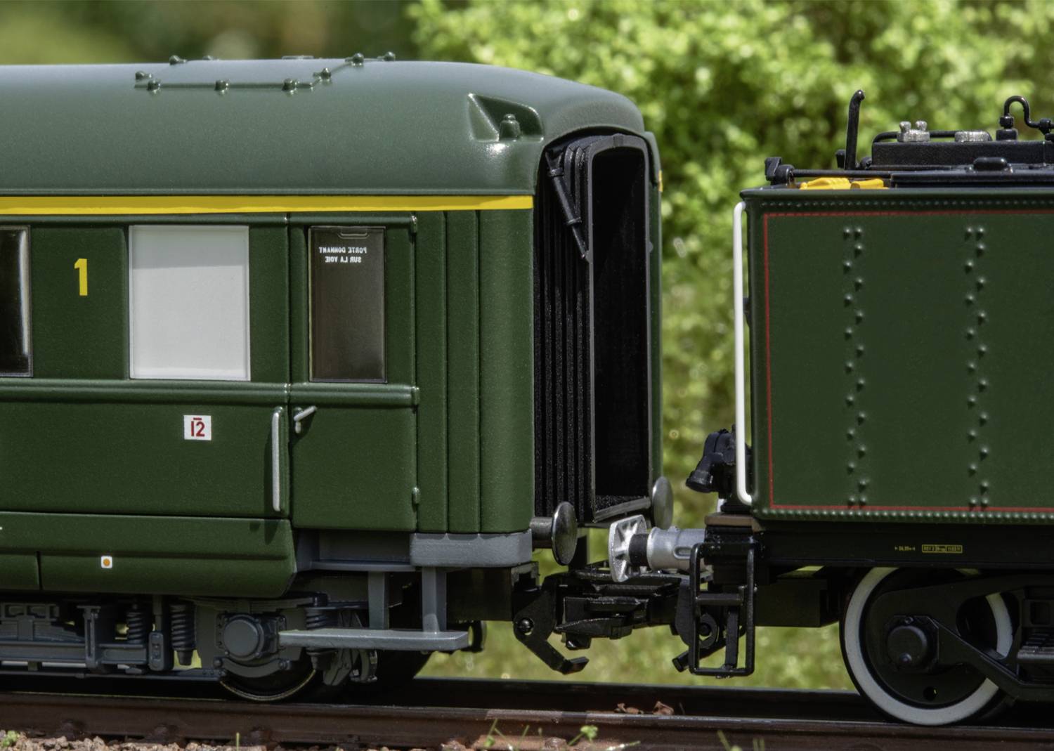 Model railway with a green passenger carriage and goods wagon coupling, on tracks outdoors. Detailed close-up of the connection.