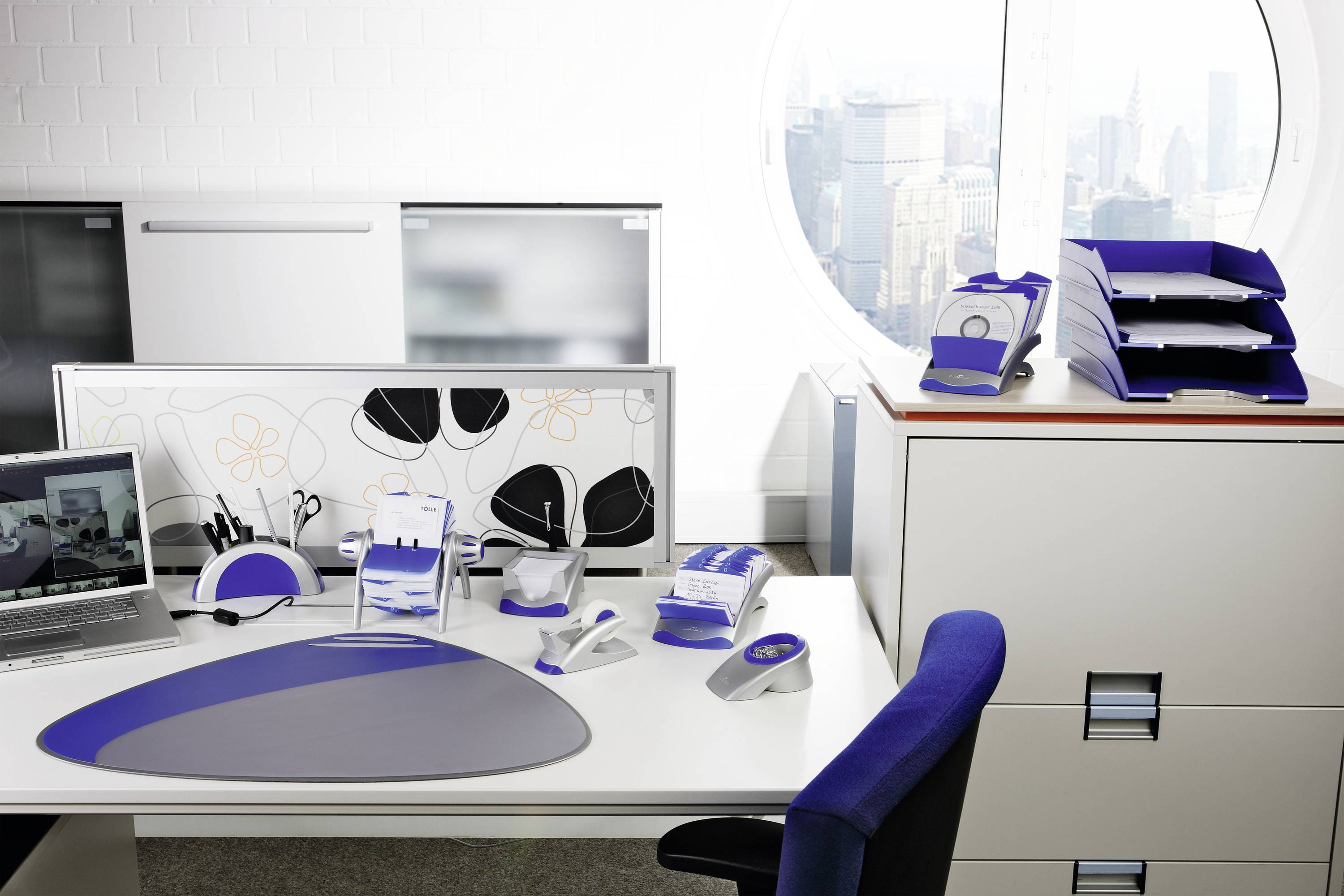 A tidy office with a desk, computer, stationery, and filing cabinets positioned in front of a round window with a city view.