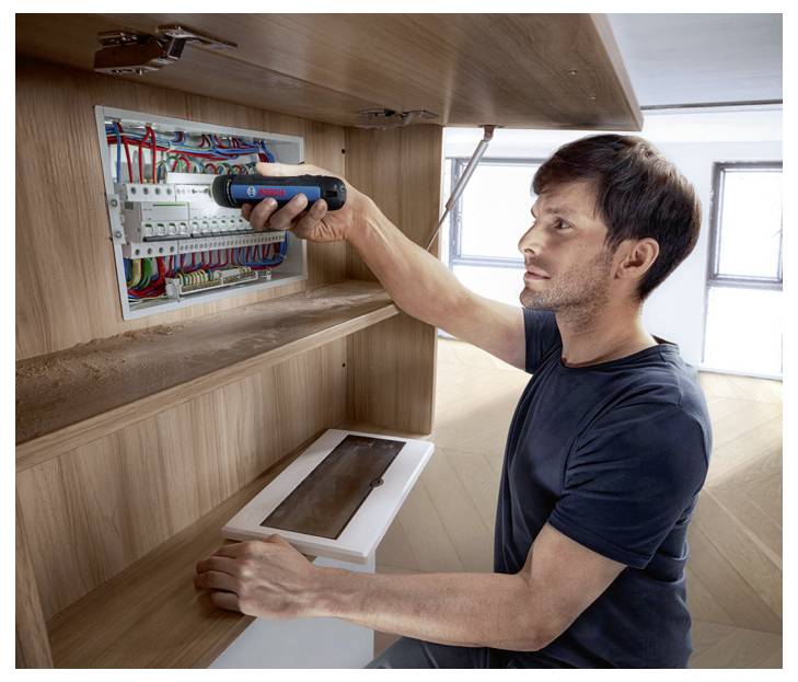 A man is repairing an electrical box with an electric screwdriver in a wooden cabinet.
