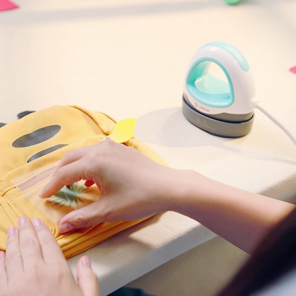 A person is ironing a small piece of fabric on a yellow bag with a sticker. A mini iron is plugged in.