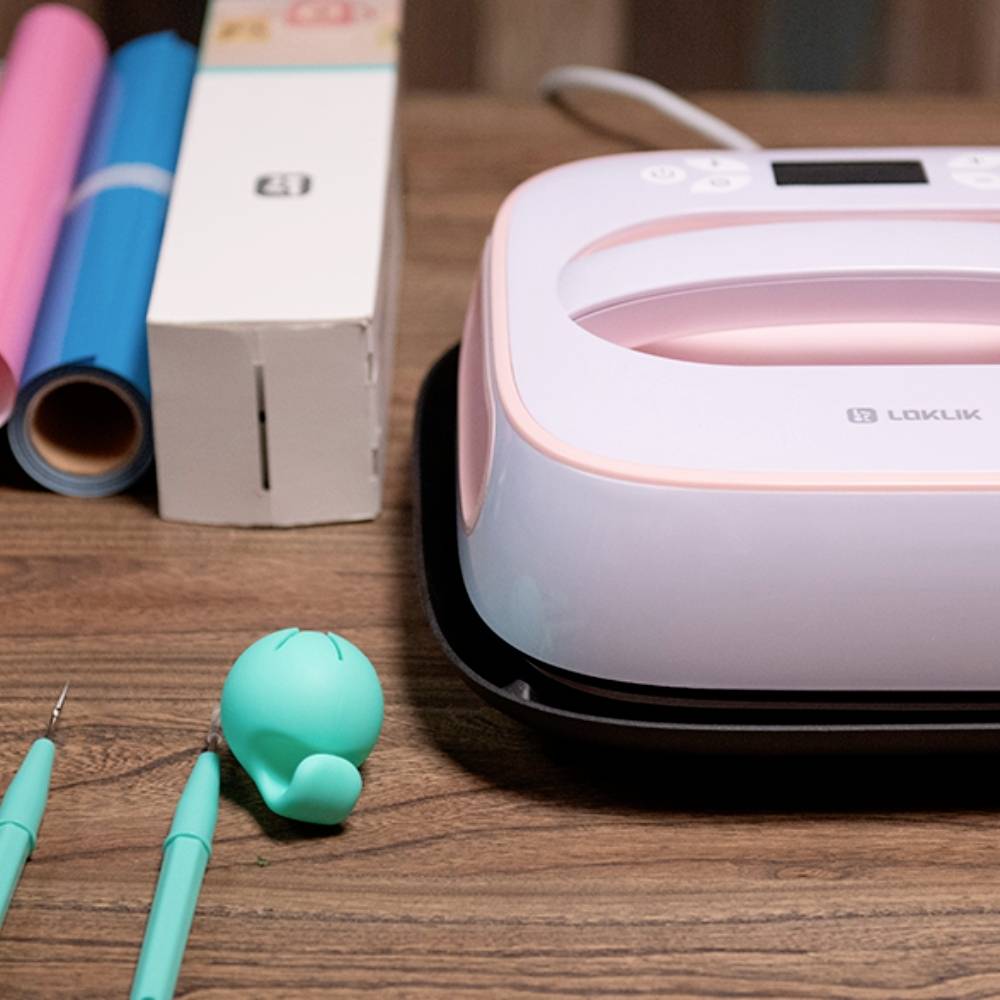 A heat transfer device sits on a table next to craft supplies, such as vinyl rolls and cutting tools, depicting a DIY creative scene.