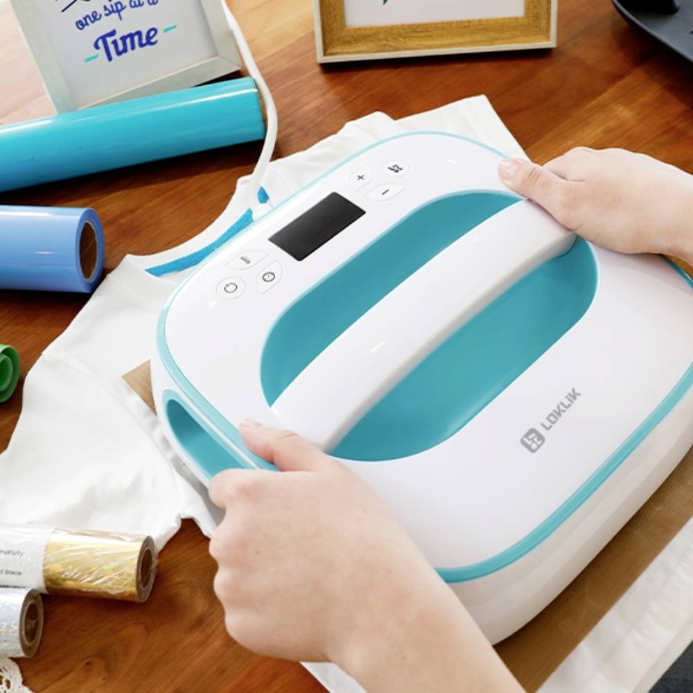 A person is operating a blue and white heat transfer machine for textile finishing. Transfer film rolls, objects, and a framed picture are located in the background.