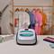 A device for printing on clothing stands on a table. In the background, colourful T-shirts hang on a clothing rail.