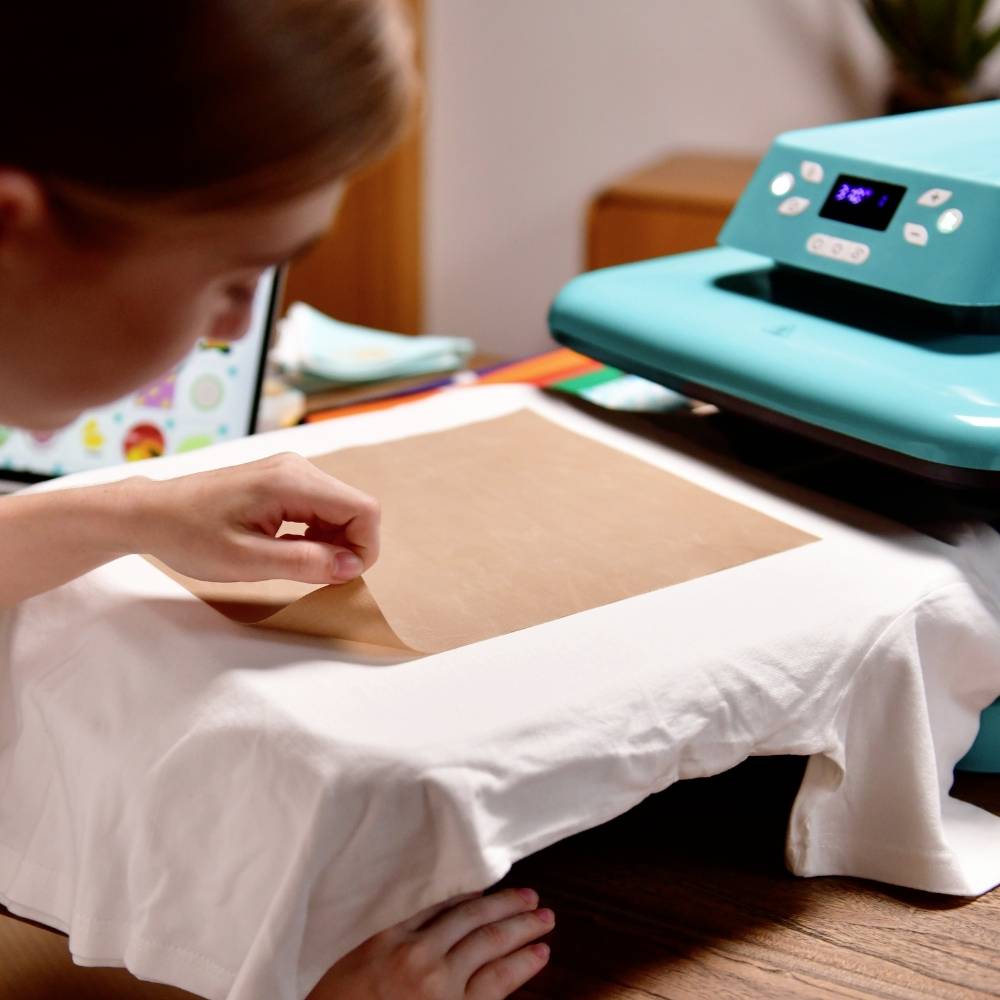 A person is using a heat press to print a design onto a white T-shirt. The transfer film is laid out ready on the fabric.