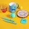 Various colourful kitchen utensils on a yellow background: mugs, glass, wooden coaster, table tennis ball, blue pens.