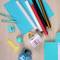 A table with two cups, two pens, a yellow spatula, a blue notepad and coloured rolls of vinyl film.