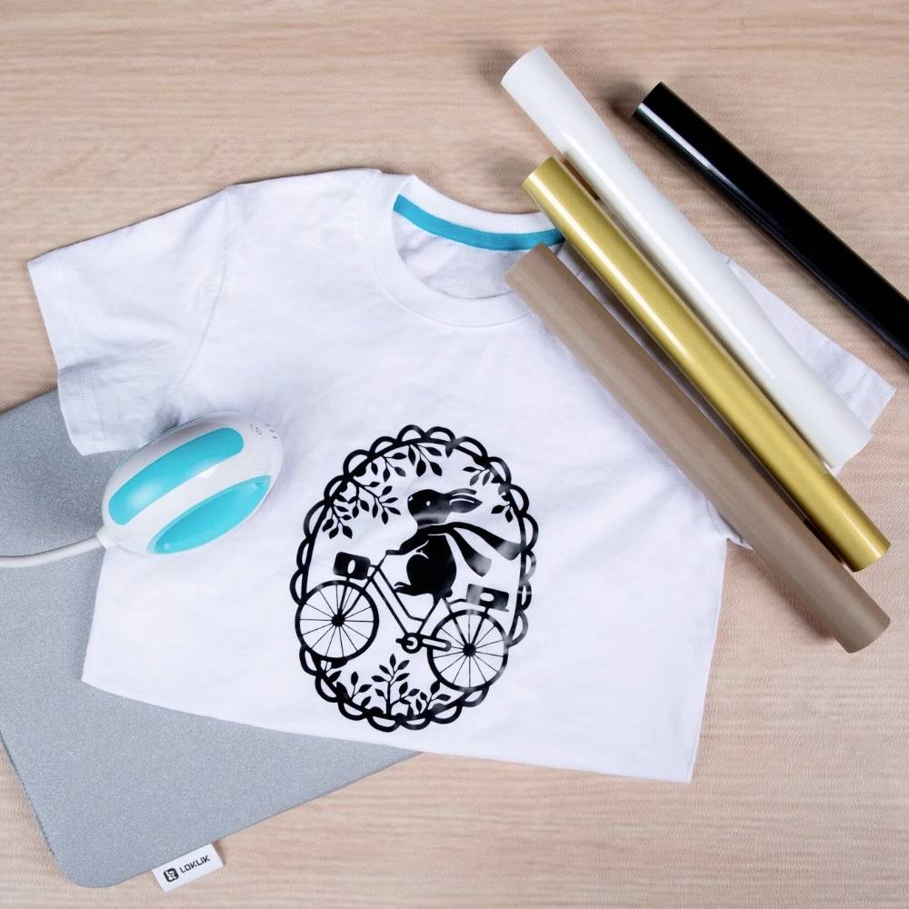 A white T-shirt with a black rabbit motif on a bicycle, next to several coloured foil rolls and a heat press on a wooden table.
