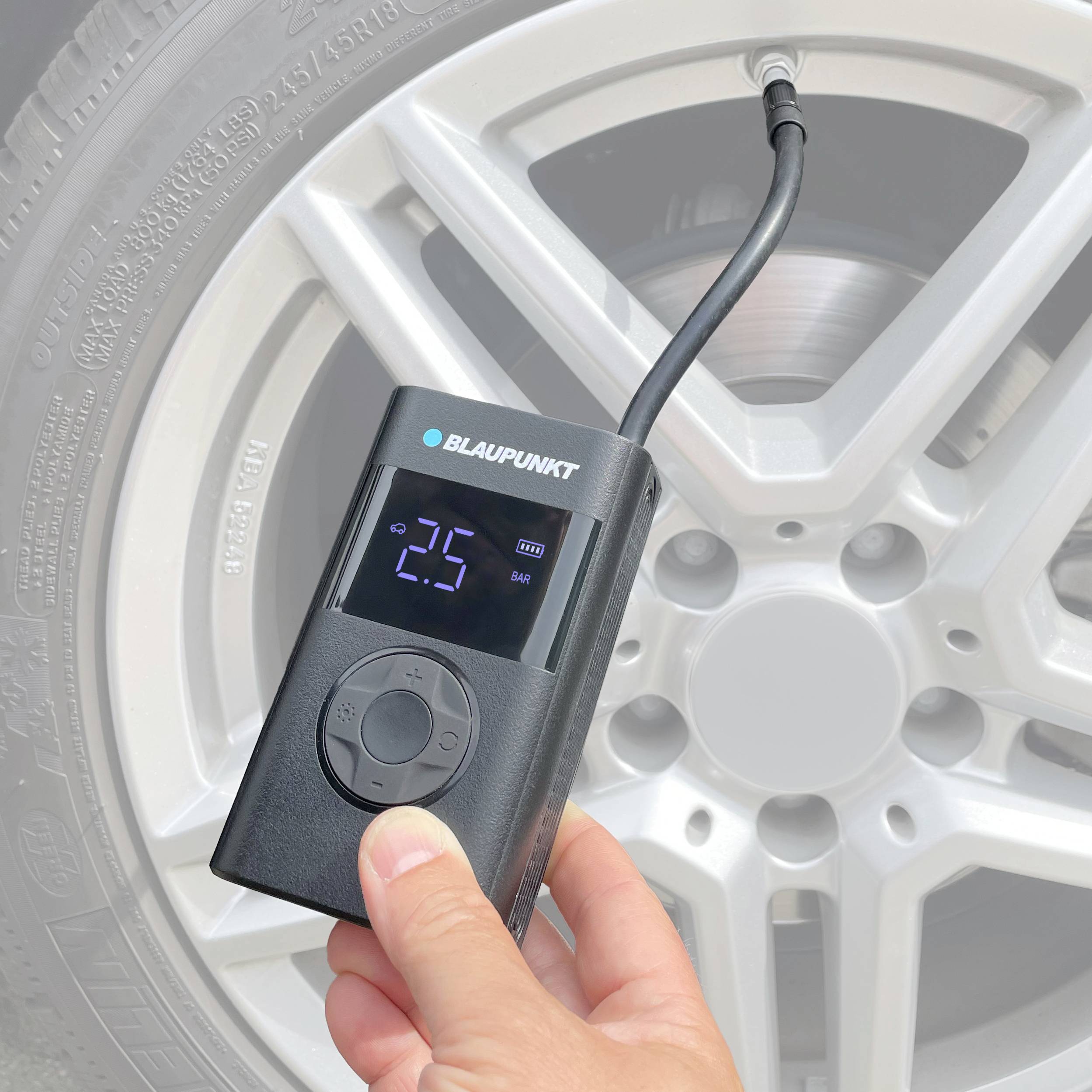 A digital tyre pressure gauge is showing '25' while connected to a car tyre. A hand is holding the device.
