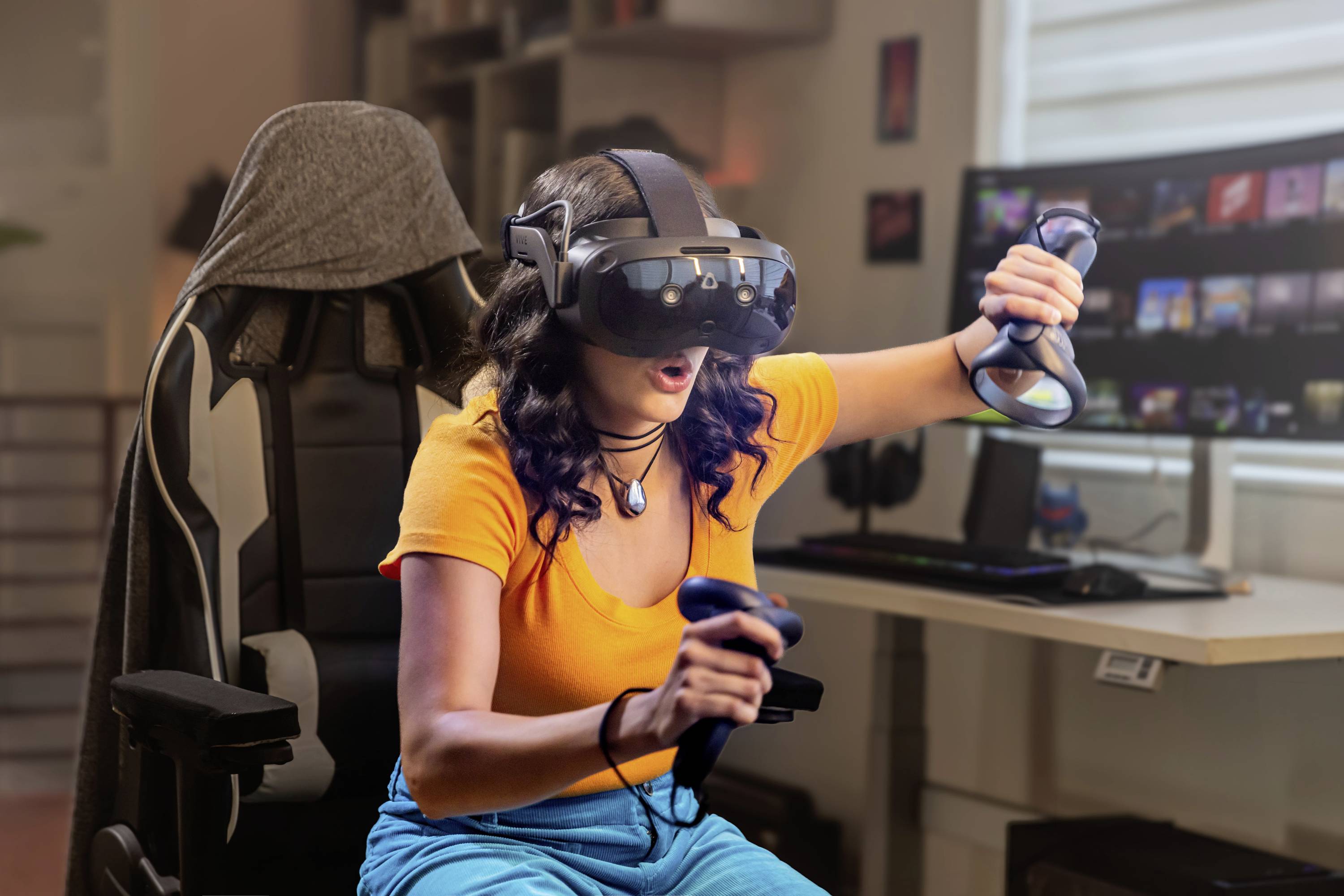 A woman is sitting in an office chair, playing a video game with a VR headset and controllers. A computer screen is visible in the background.