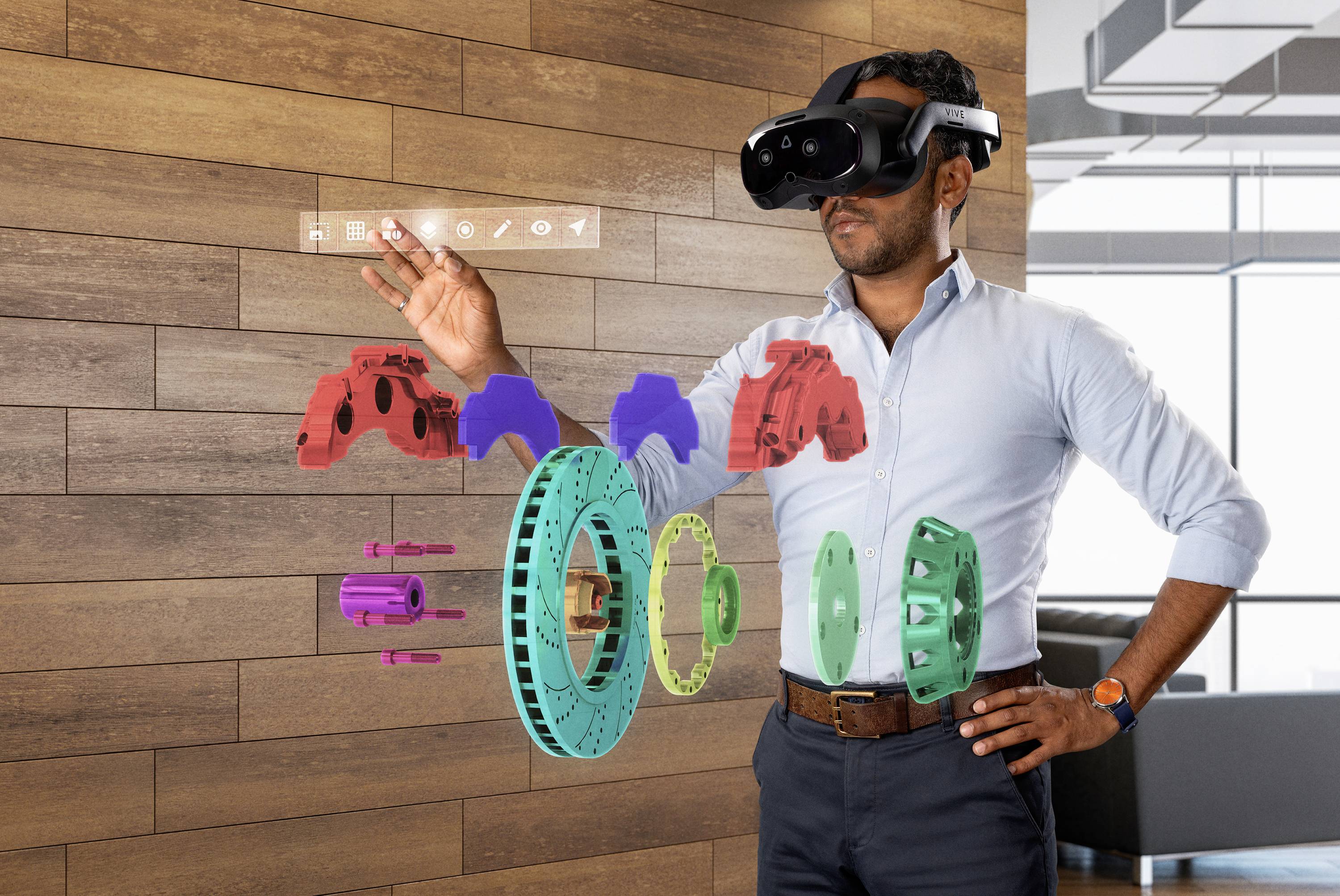 A man wearing a virtual reality headset interacts with floating 3D renderings of car parts in a modern office.