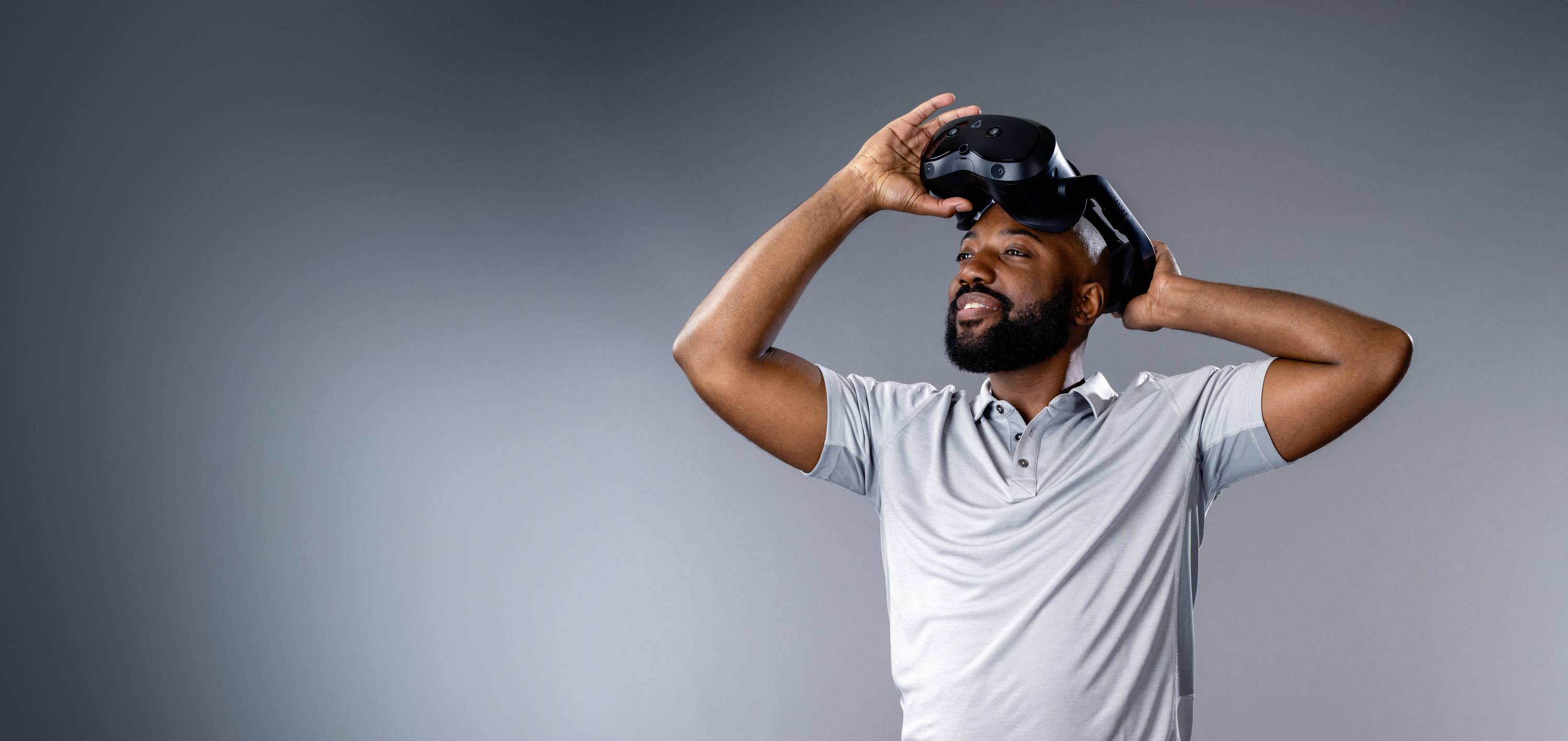 A man is wearing a virtual reality headset and looking upwards. The background is a uniform grey.