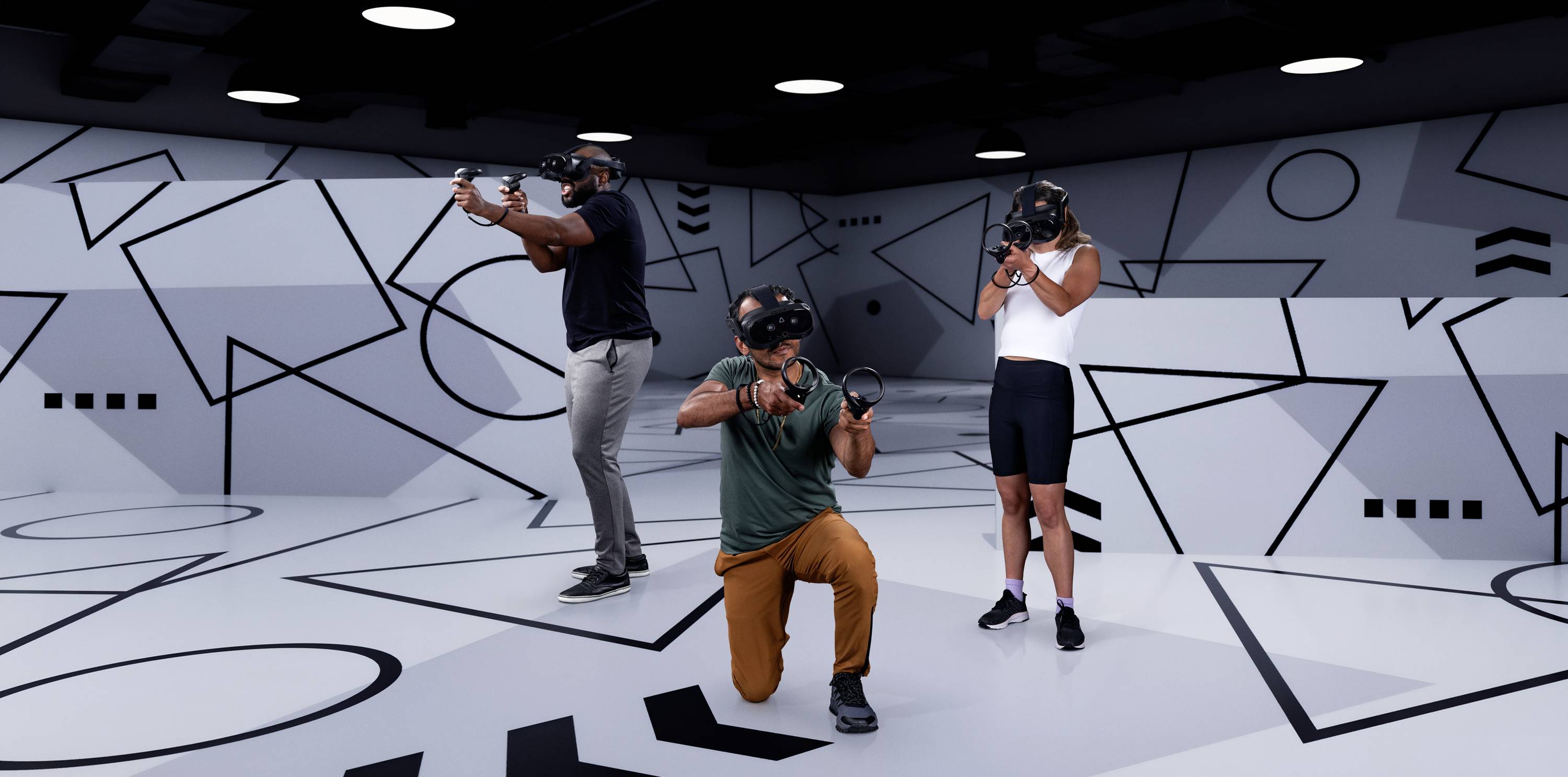 Three people are wearing VR headsets and standing in a room with geometric graphics on the walls, while using VR controllers.