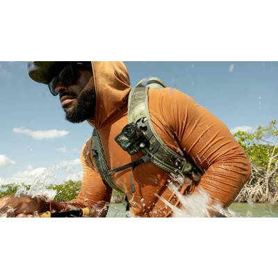 Person in an orange hoodie, wearing sunglasses and carrying a rucksack, wades through shallow water. The sky is clear, with mangroves in the background.