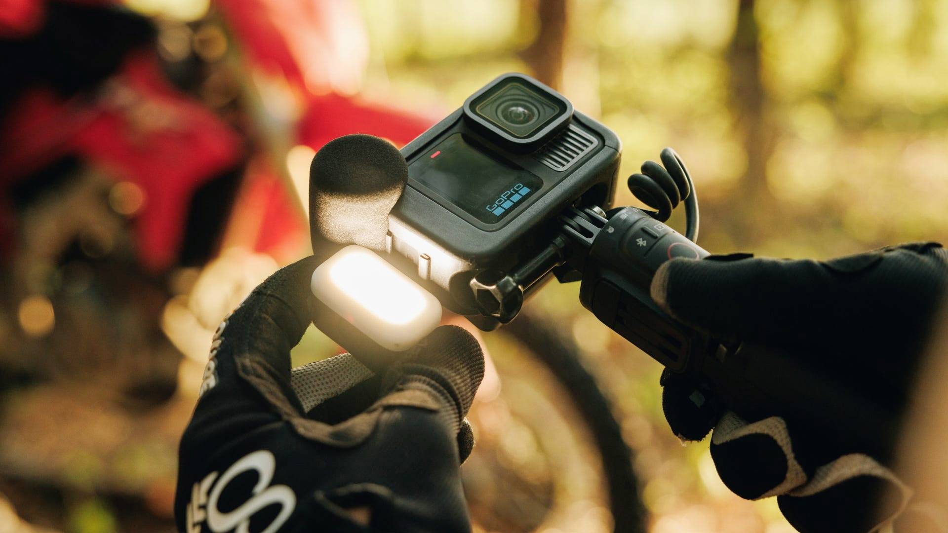 A person wearing black gloves is holding an action camera and a microphone. In the background, a blurred red motorbike can be seen.