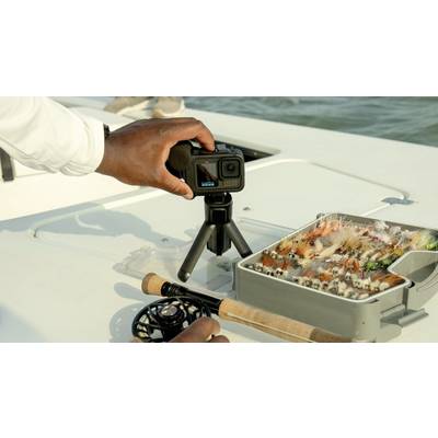A person is preparing a camera on a boat table. Next to the camera is an open box with fishing flies.
