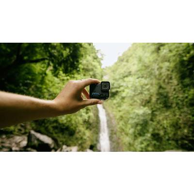 A hand holds an action camera in front of a blurred background featuring a waterfall and dense forest.