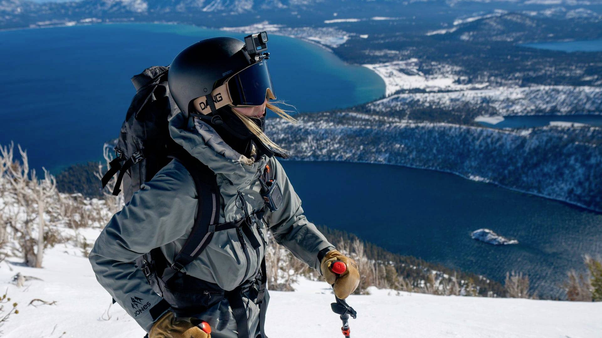 A person in ski gear stands on a snow-covered slope, looking out over a vast lake and forested landscape in the background.