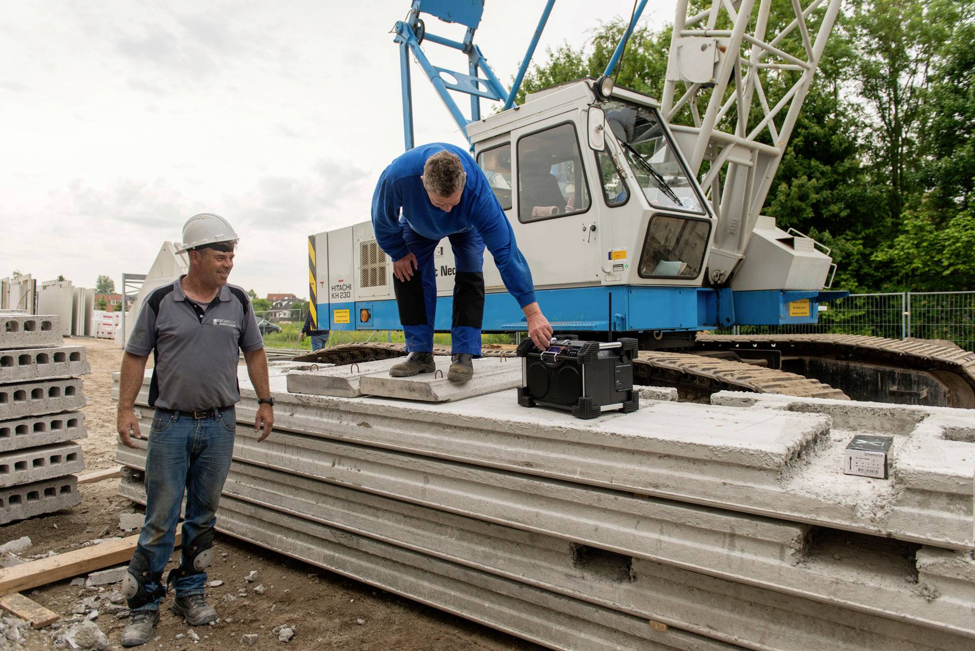 A construction worker is lifting a Bluetooth speaker on concrete slabs while another worker wearing a hard hat stands nearby. A crane is in the background.