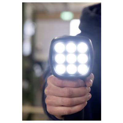 Close-up of a hand holding a bright square LED flashlight or work light, shining directly toward the camera indoors.