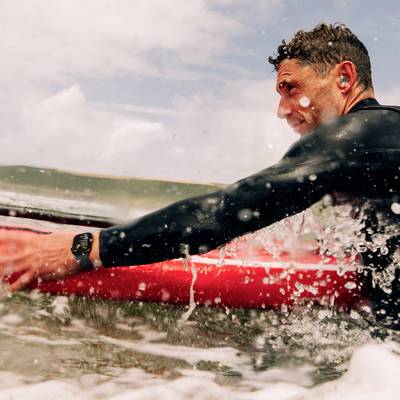 A surfer is wearing a wetsuit and is lying on a surfboard in choppy water. He appears to be preparing to paddle out.