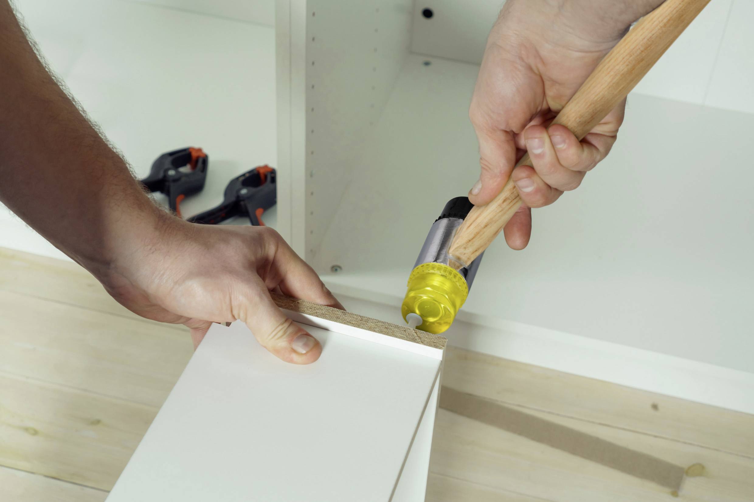 A person is assembling a white piece of furniture and hammering a nail into the edge of a board. Tools are laid out ready.