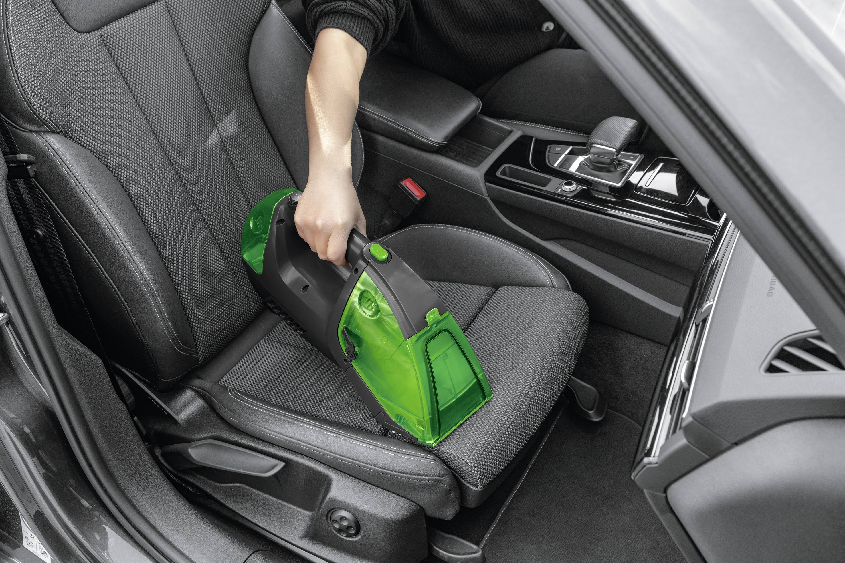 A person is cleaning the driver's seat of a car with a green handheld vacuum cleaner. The interior is modern and clean.