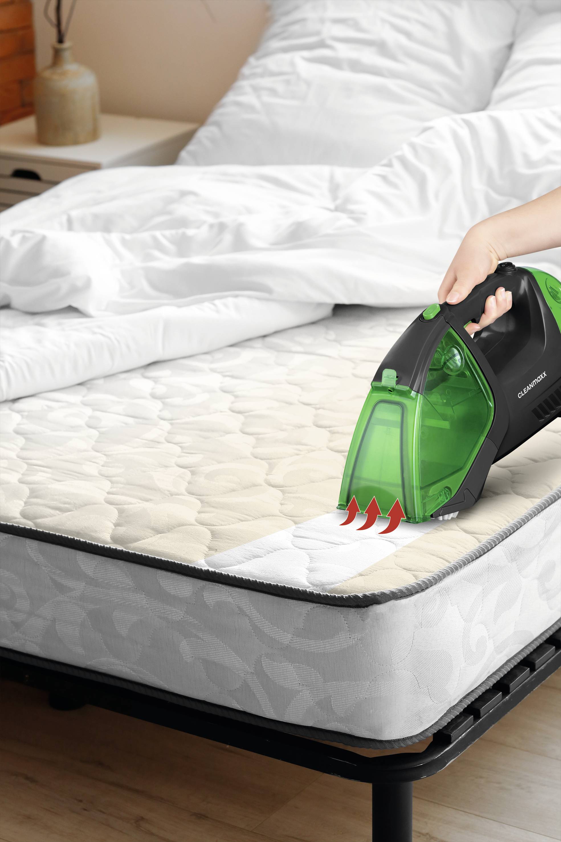 A person is cleaning a mattress with a green handheld vacuum cleaner. Bedding and a bedside table are visible in the background.