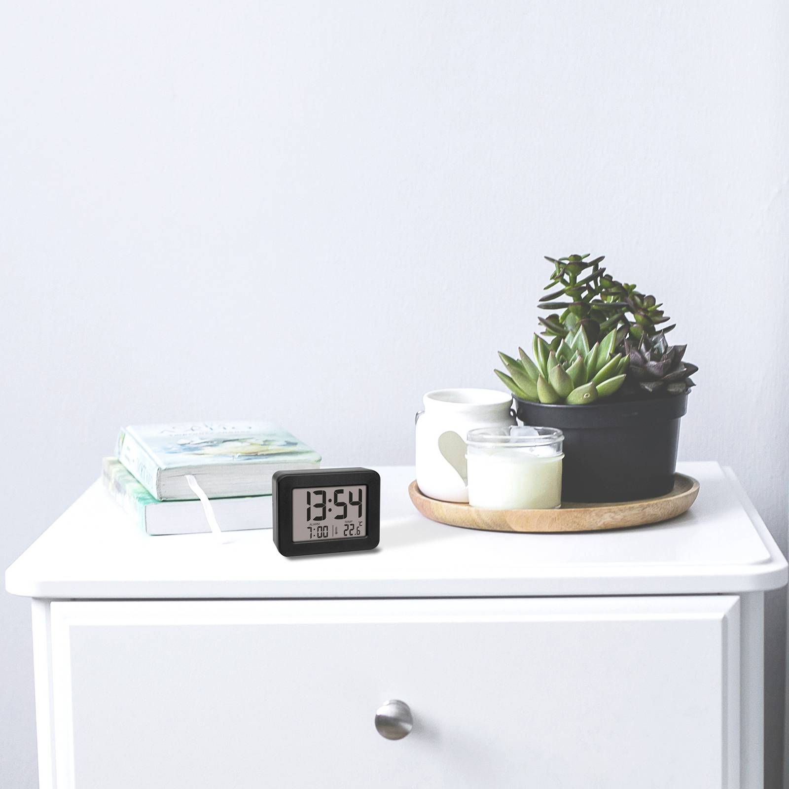 A bedside table with a digital clock showing 13:25, candles on a tray and a book. Plants are standing in the background.