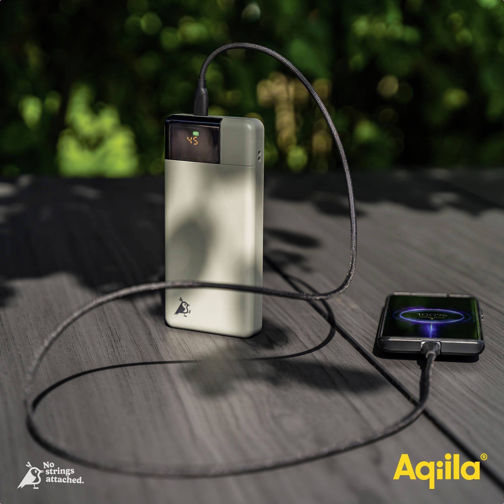 A portable charger with cables is charging a smartphone on a table outdoors. The brand 'Aqiila' is visible in the background.