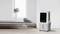 An air cooler stands in a modern, minimalist room with wooden flooring. Next to the cooler, grey cushions are placed on a bench.