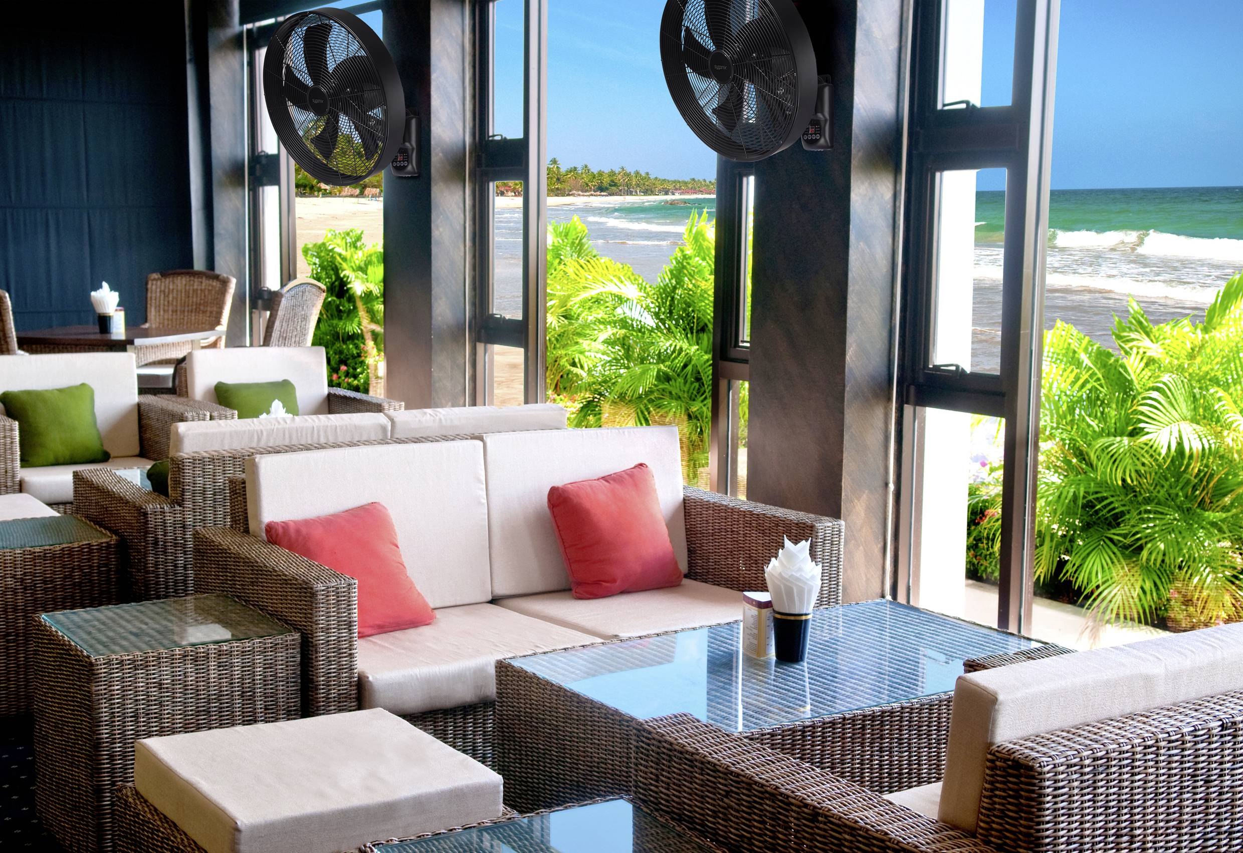 A stylish beach lounge with wicker furniture and colourful cushions. Large windows offer a view of palm trees and a sunny, blue sea.