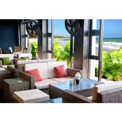 A stylish beach lounge with wicker furniture and colourful cushions. Large windows offer a view of palm trees and a sunny, blue sea.