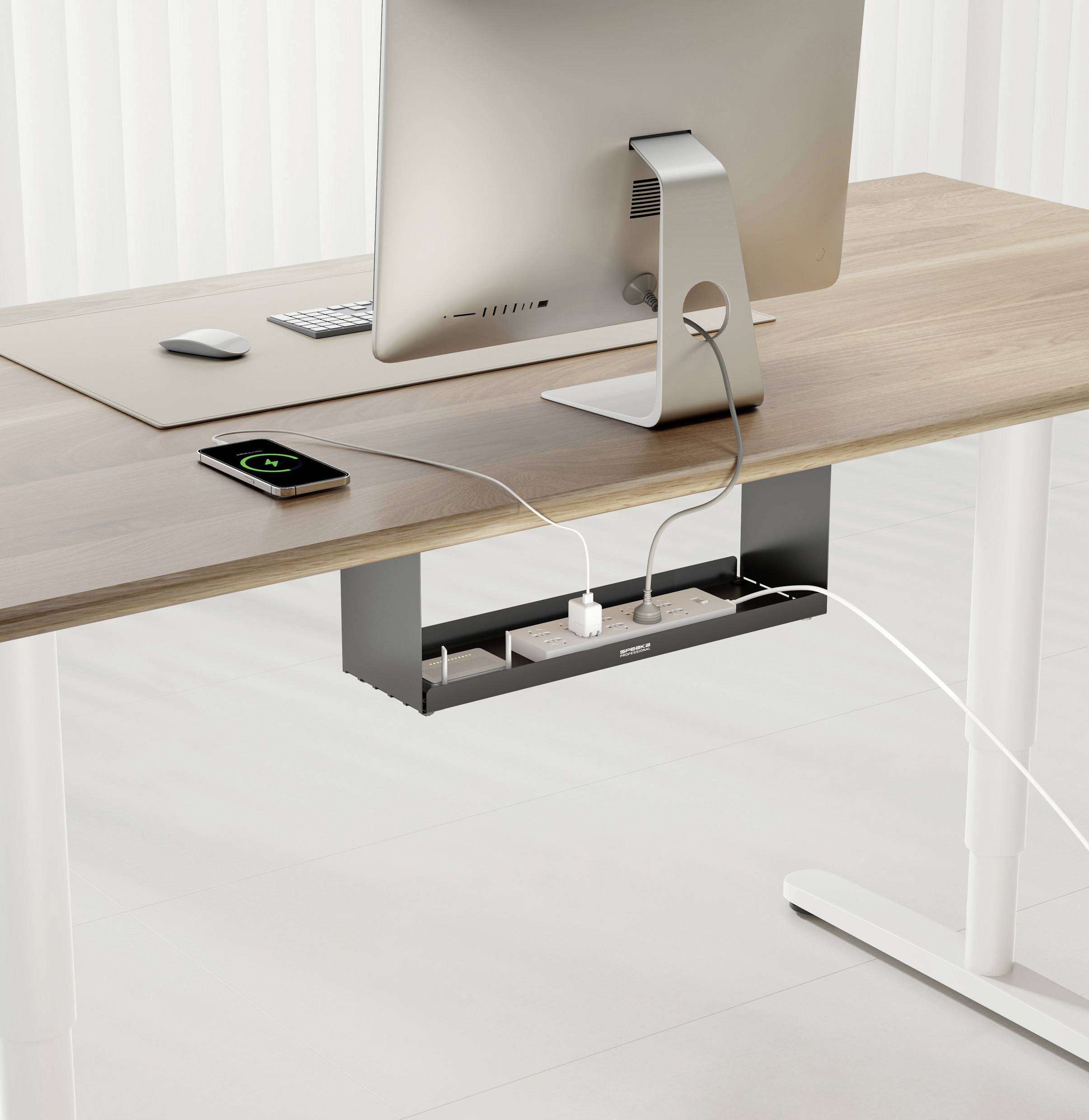 A modern desk with a monitor and a smartphone being wirelessly charged. A multi-socket extension lead is mounted beneath the table.
