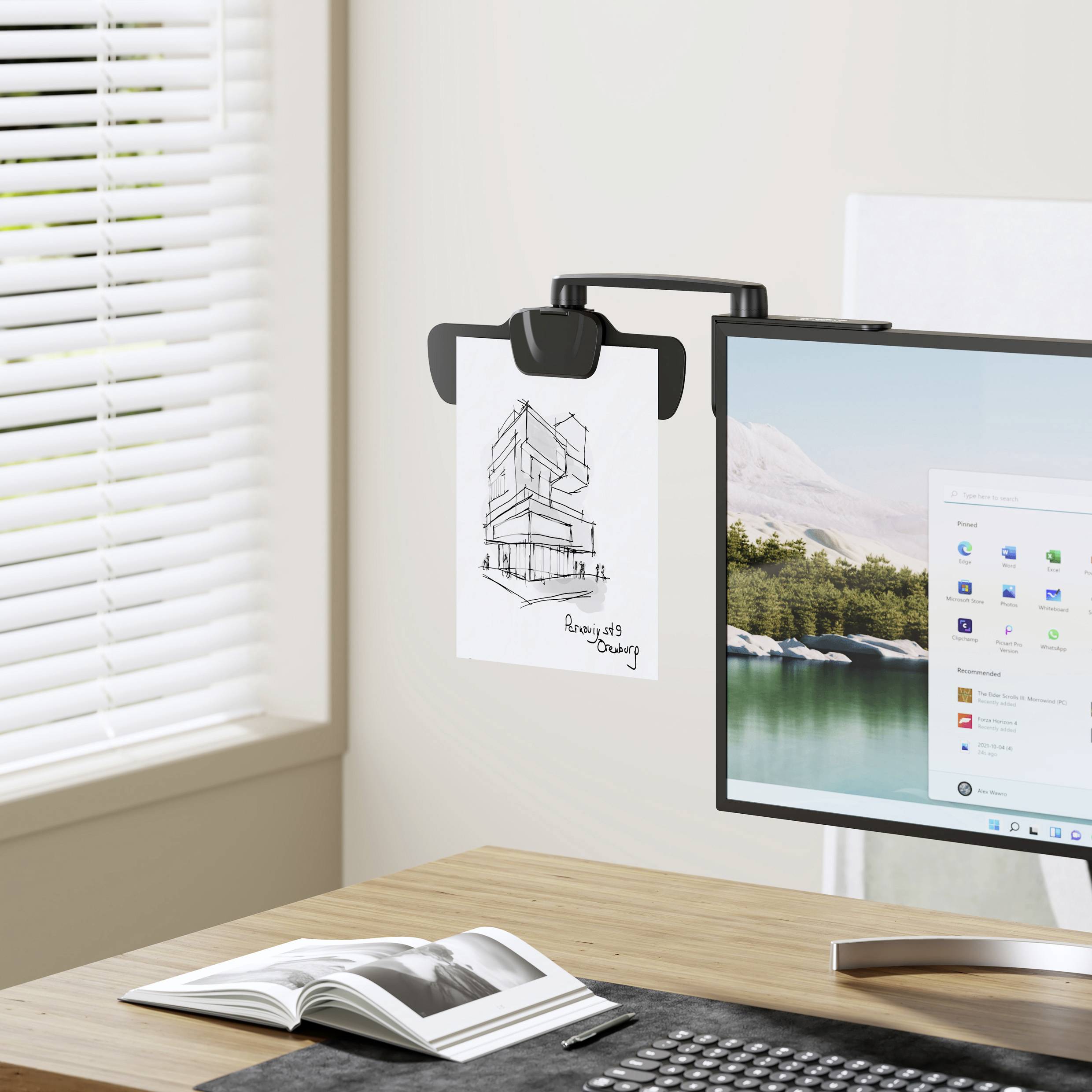 A computer monitor displays the Windows user interface. A drawing of a modern building is hanging beside the monitor. A window is visible in the background.