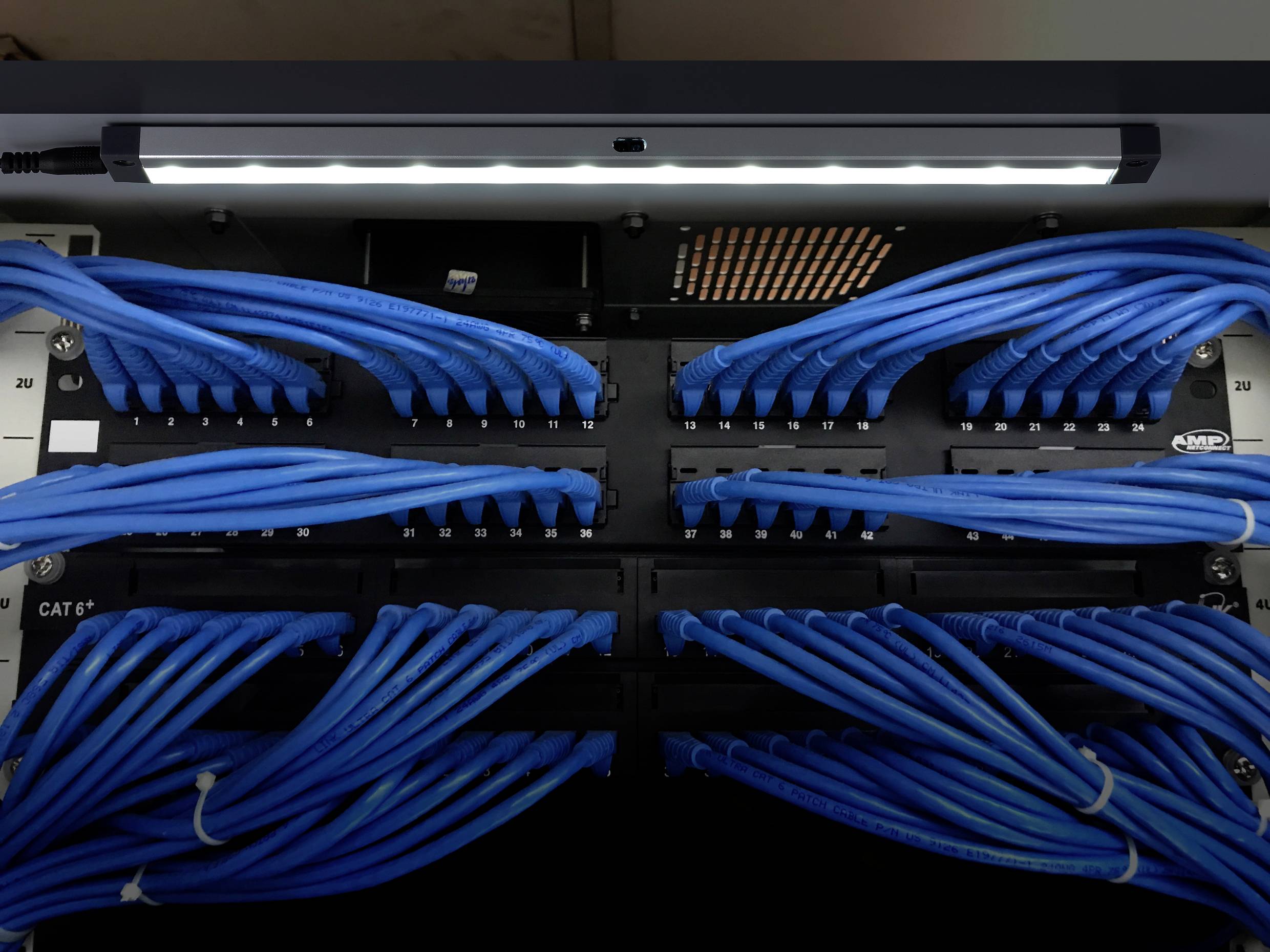 A rack-mounted network patch panel with blue Ethernet cables connected in rows, illuminated by an overhead light.