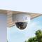 Surveillance camera mounted under a roof, pointing downwards, with a view of blue sky and green trees in the background.