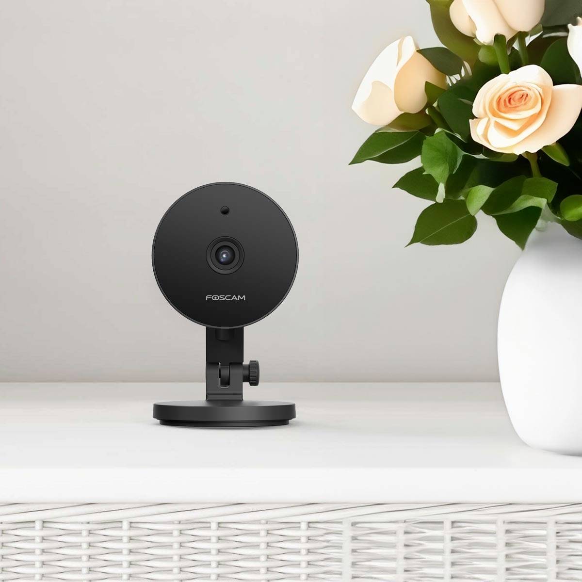 A black Foscam surveillance camera sits on a white table beside a white ceramic vase with pink flowers.