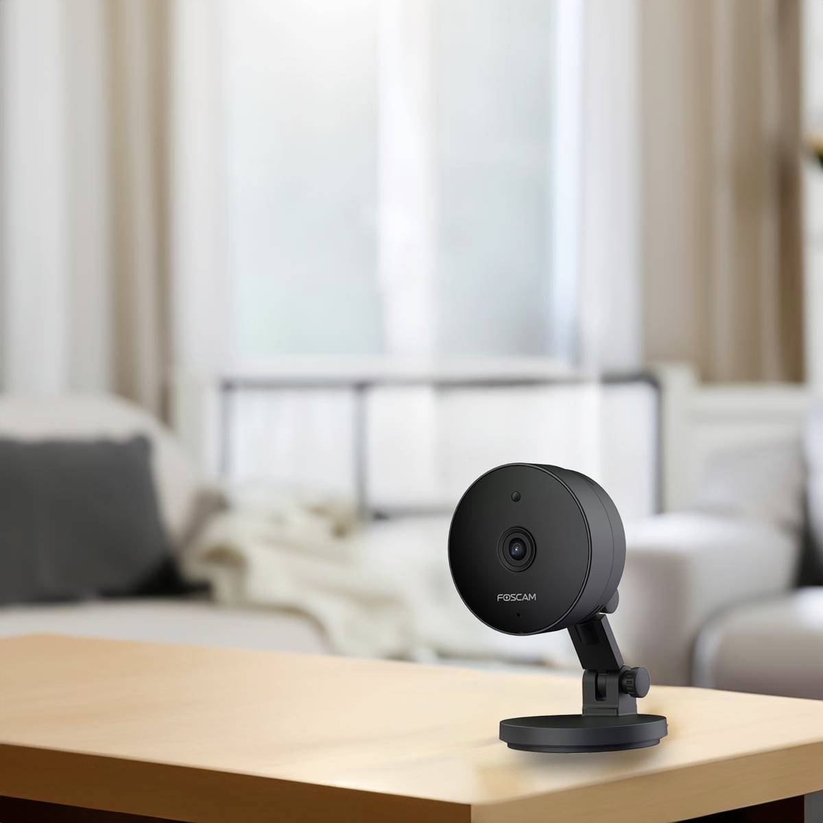 A surveillance camera on a table, with a blurry living room with a sofa and cushions in the background. The camera is facing forward.