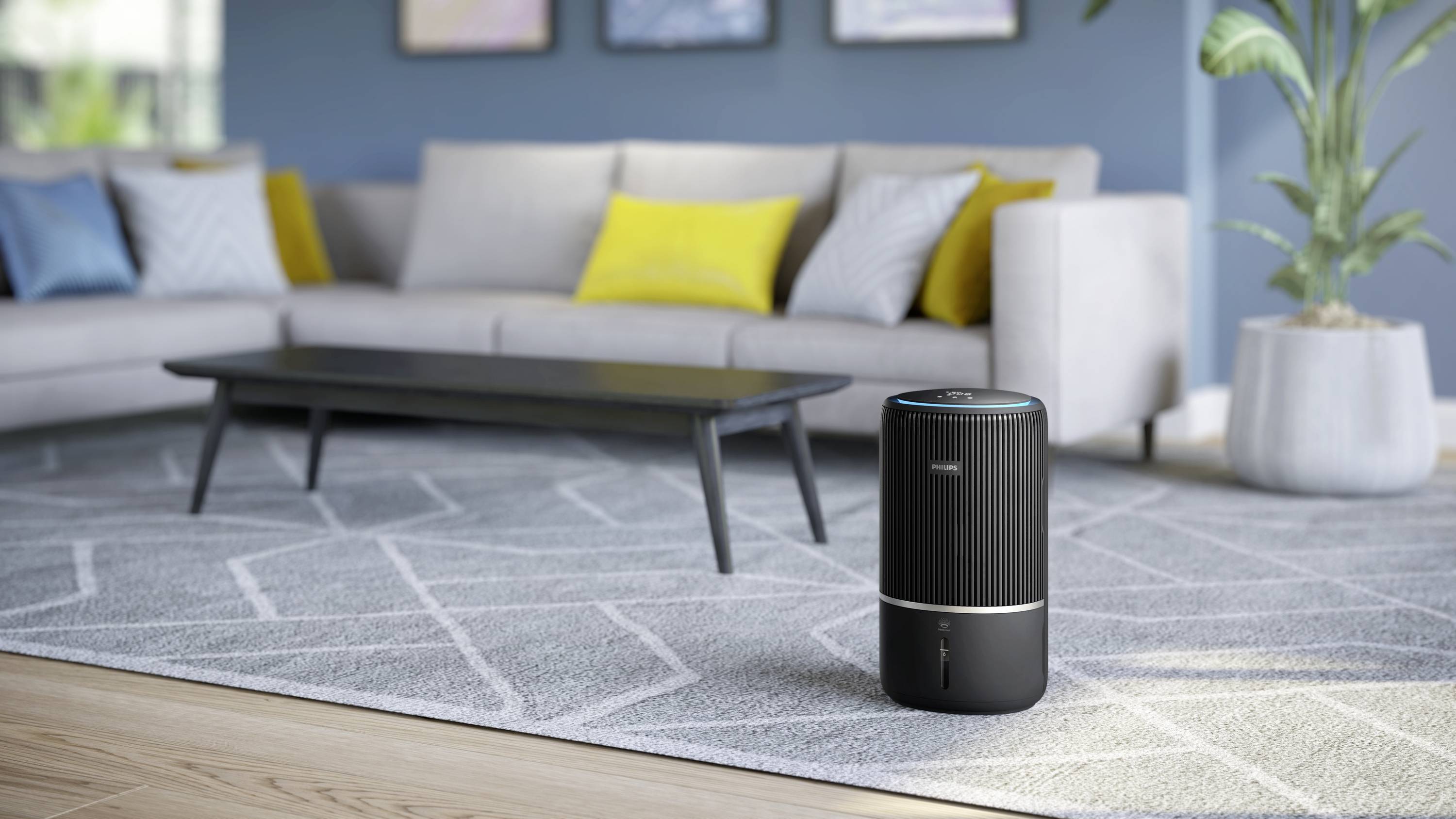 An air purifier stands on a carpet in a modern living room with a grey sofa, yellow cushions, and a low table.