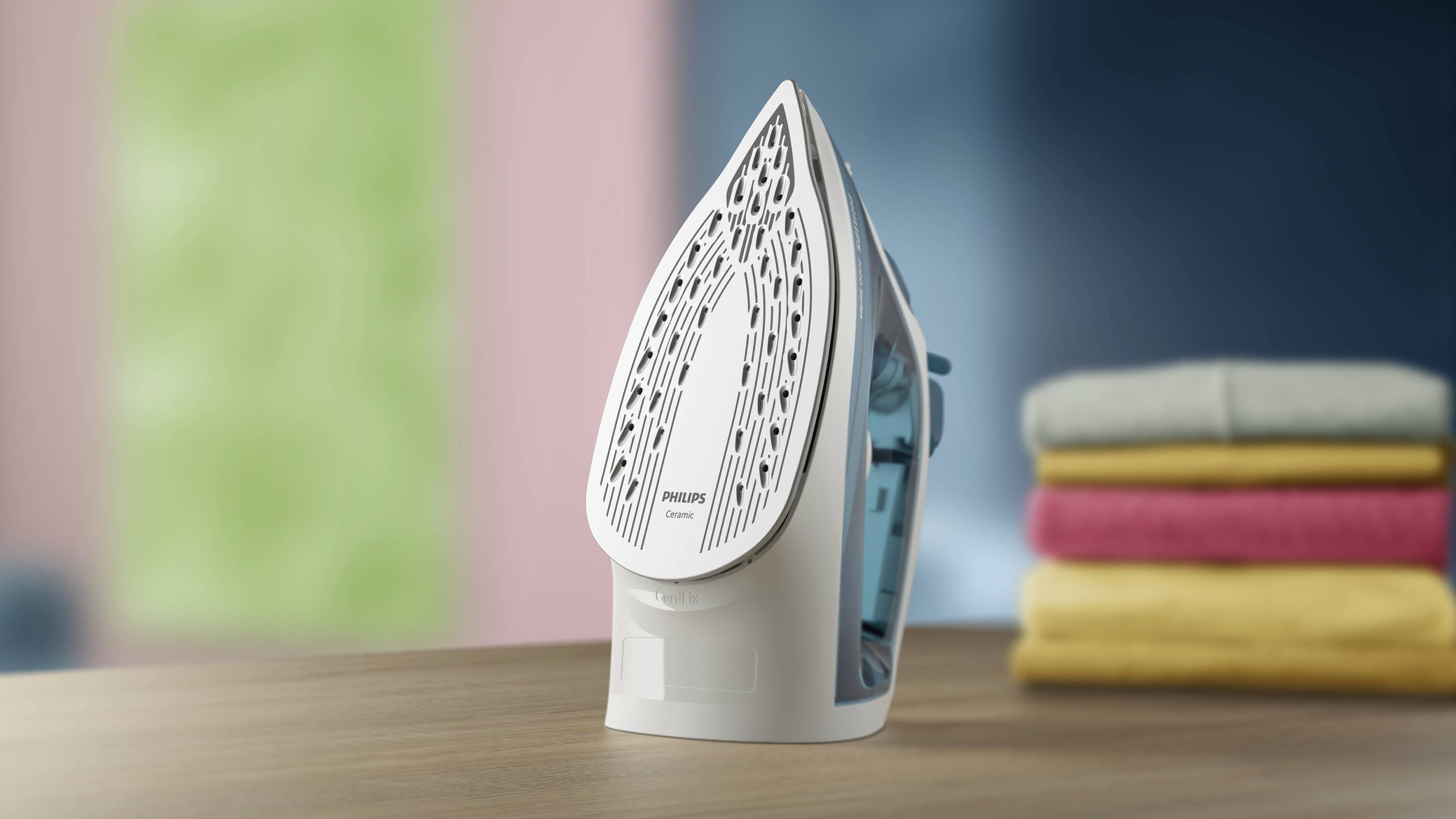 An iron is standing on a table next to a stack of folded clothing. Blurred colours are visible in the background.