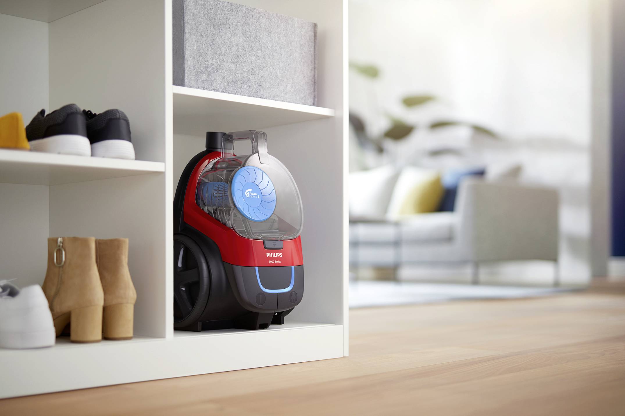 A red vacuum cleaner sits on a shelf with shoes, with a living room in the background; clean and tidy.