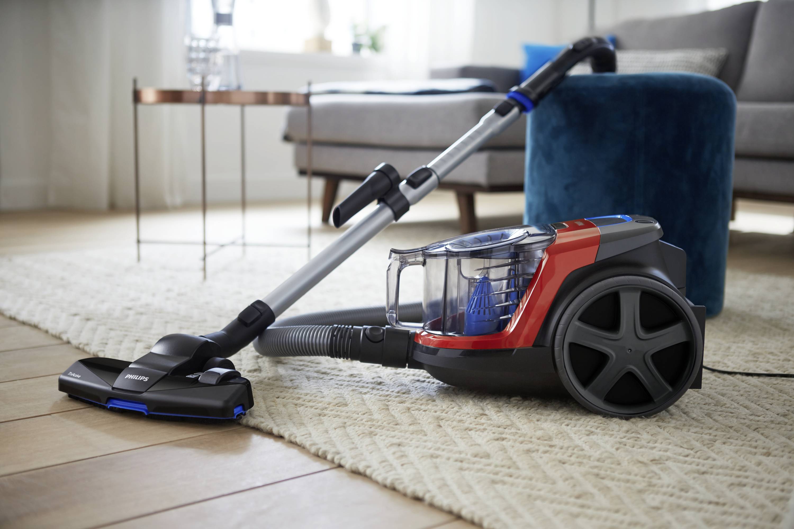 A vacuum cleaner on a carpet in a living room. A sofa and a small table are visible in the background.