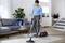 A woman is hoovering in the living room. She is standing on a light-coloured carpet, next to a grey sofa and round table.